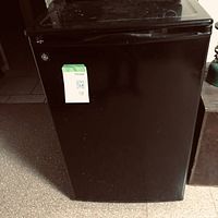 Front closed view of a black compact bar fridge with slightly reflective surface and a label on the top left corner.