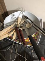 Various BBQ skewers with wooden handles, metal tongs, wooden spoons, and plastic mixing bowl shown.