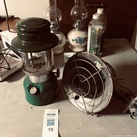 Green Coleman camping lantern seen next to propane heater and some cleaning products on a concrete floor.