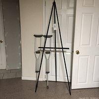 Full view of two small adult crutches and a black plastic artist easel standing upright against a white door in a room.