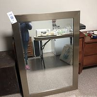 Large rectangular wall mirror leaning against a wall showing a brushed metallic frame and clear reflective surface.