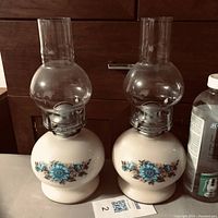 Two vintage oil lamps with white glass bases decorated with blue and pink flowers, clear glass chimneys, and metal burners.