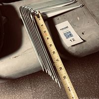 Photo showing nine white steel shelf brackets stacked with a measuring tape indicating the length of approximately 8 inches.