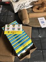 First photo showing one open box containing multiple vintage match packets with distinctive provincial shield designs and colorful text on top, surrounded by wooden and metal kitchen utensils.