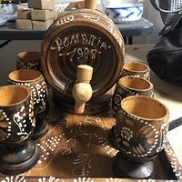 Close-up of wooden keg barrel with spigot and markings 'Romania 1998' surrounded by six decorated wooden cups on a matching tray.