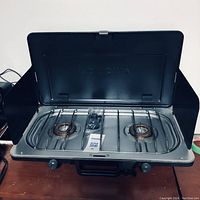 Front open view of Woods 2 burner camp stove showing burners, grates, control knobs, and lid with wind guards.