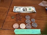 Photo showing one 1976 $2 bill and seven coins arranged on a wooden table with a green label specifying years and types of coins.