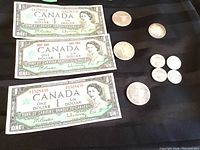 Three Canadian Centennial one dollar bills from 1967 together with various coins including dollars, a 50 cent piece, and quarters displayed on black fabric.