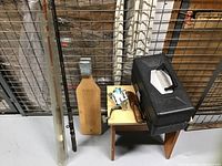Fishing gear assembled near metal grid behind, includes black plastic tackle box, two fishing rods, fish scaler board, and small wooden bench