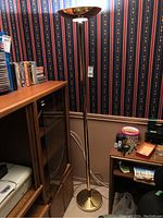 Full view of brass torchiere floor lamp standing beside wooden furniture, showing overall lamp shape, design, and height.