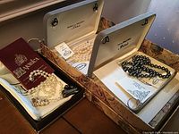 Three sets of Majorica pearls in open presentation boxes on wooden surface. Includes white pearl necklaces and earrings, black/grey pearl necklace and ring.