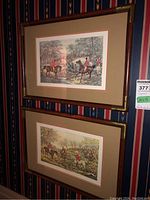 Two framed prints vertically hung on a patterned wall showing fox hunting scenes with riders and dogs. Frames are brown wood with gold corners and beige mats with a red border inside.
