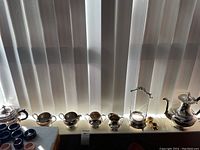 Wide shot showing the full set including pots, cream and sugar containers, pickle caster, sugar tongs, crumb tray, and brush all reflective silver plate items on a windowsill.