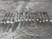 Overview of full set of fifteen sterling silver souvenir spoons arranged in a line, showing various handle designs and markings.