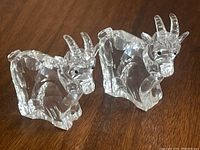 Both Swarovski crystal goat figurines shown side-by-side on a wooden surface, one with a broken horn.