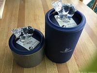 Two Swarovski bear figurines placed on their original blue cylindrical boxes, clear faceted with gold fish in mouth, on a wooden table.