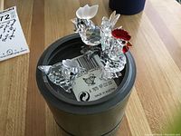 Three Swarovski crystal rooster and chick figurines displayed on a cylindrical box with a barcode label visible. Clear multifaceted crystal with distinctive frosted combs and a red comb/wattles on the largest rooster.