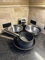 Photo showing two stainless steel mixing bowls, one black saucepan with lid, one black frying pan, and a box grater on kitchen counter.