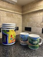 Photo showing colorful Chaleur Starbucks coffee tin with metal latch lid and stacked coasters on top, alongside two ceramic mugs with scenic landscape designs.