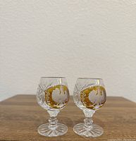 Two small vintage crystal glasses with amber and white eagle detailing on the front, sitting on wooden surface against plain wall.