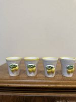 Four white ceramic shot glasses with yellow lemon and green leaf designs on the front, arranged side by side.