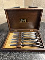 Full set of eight steel steak knives neatly arranged inside a wooden box with yellow felt lining.