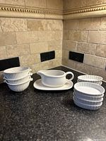 Vintage white ceramic crème brûlée set with five round ramekins, two oval ramekins, and sauce boat with underplate shown on tiled counter.