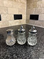 Three vintage cut glass salt and pepper shakers on a dark countertop with beige stone tiled backsplash.