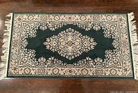 Full view of the rectangular vintage rug with green geometric central medallion and floral border, fringed edges visible.