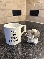 Photo showing white ceramic mug with black text 'FOR MY BEST FRIEND' beside small ceramic angel figurine on black countertop