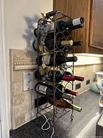 Side view of vintage metal standing wine rack holding 12 wine bottles, decorative grape leaves and grapes detail visible, placed against kitchen backsplash.