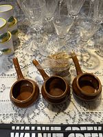 Three brown glazed ceramic bowls with handles arranged on a lace tablecloth, with various glassware in background.