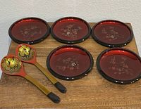 Five lacquered plates laid out individually with two lacquered spoons showing detailed floral designs and gold accents
