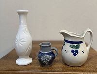Three pitchers displayed side by side on wooden surface: tall white milk glass, small blue ceramic, and white porcelain with painted grape motif.