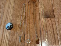 Five pieces of jewelry displayed on wood floor surface: one round marbled pendant or pin, and four necklaces with various pendants including one gold 'R' initial pendant with clear gemstone.