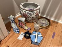 Photo showing all items in the lot arranged on a wooden floor: two ceramic shoe figurines, large floral planter, two candle holders, stained glass box, and framed textile.