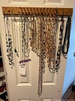 Photo showing multiple necklaces hanging on wooden pegs on a door, including beaded strands in orange, cream, turquoise, and silver tones, as well as black chain necklaces.
