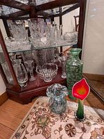 Collection of clear crystal cut glass bowls and vases inside display cabinet and three colorful blown glass vases and candlestick on floor in front of cabinet.
