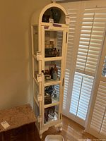 Full view of tall painted wood curio cabinet with framed glass door and glass sides, placed near window with shutters. Cabinet stands on four legs.
