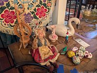Full lot view on wooden table with wool tapestry, showing spinning wheel, doll, eggs, plastic swan, pottery vase, figurines, and lizard