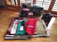 Holiday-themed linens including embroidered and crocheted placemats and table runners in red, green, and plaid patterns stacked on floor with boxes and pillows behind.