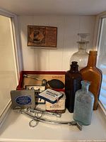 Single image showing a group of vintage medical bottles, pharmaceutical tins, metal medical tools, and a boxed DeVilbiss medical bulb syringe tool.