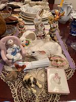 Overview of the twelve figurines displayed on a lace cloth showing various pieces including different animal and human figures, decorative dish, and a porcelain plaque.