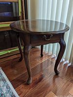 Oval wooden side table with curved legs and a central dovetail drawer with metal handle, showing surface scratches.