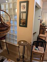 Wood framed mirror mounted on wall with artwork and mirror panels, mosaic plant stand on floor below it, glass block wall to left, chairs and other home furnishings in background.