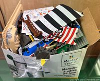 Overhead view of box containing assorted LEGO pieces including fabric flags, tan hull panel and grey plates
