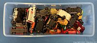 Top-down view of approximately 20 small die-cast toy vehicles in a clear plastic bin, showing a mix of cars, trucks, and other models including an ambulance and school bus.