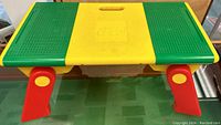 Green and yellow Lego builder table with red foldable legs and a handle slot in the center, showing front view