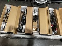Four cardboard boxes each containing slim, long metal and plastic display lighting fixtures with attached black power cords, laid side-by-side on the floor.