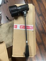 Photo of one Skandalight exhibit display stand light laid on a cardboard box showing cylindrical housing with heat sink fins and adjustable mounting arm.